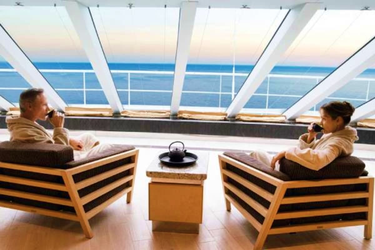 Passengers relaxing in lounge chairs on a cruise ship observation deck with ocean and sky views through large windows