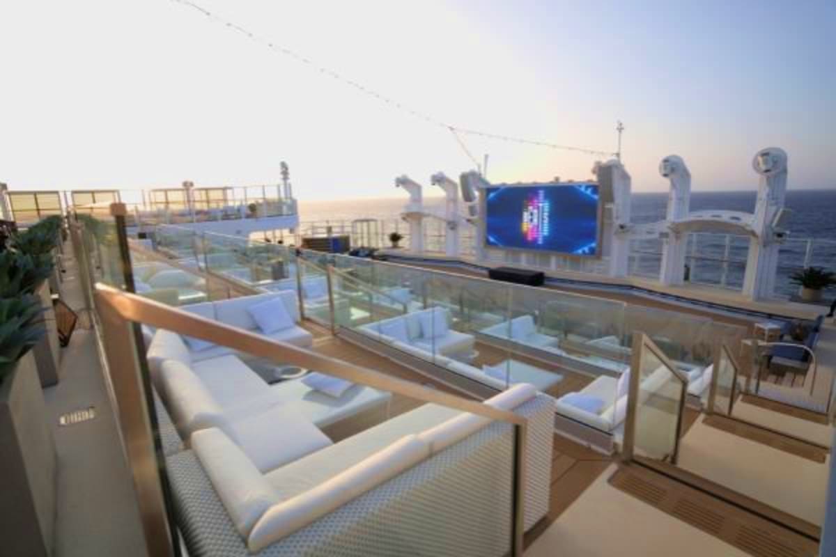 Luxury cruise ship deck with white lounge seating, glass railings, digital display screen, and ocean horizon at sunset