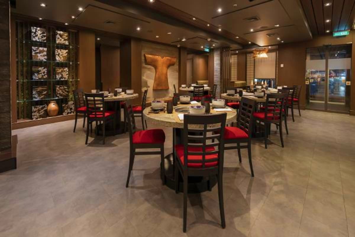 Modern cruise ship dining room with round tables, black chairs with red cushions, warm lighting, and contemporary Asian-inspi