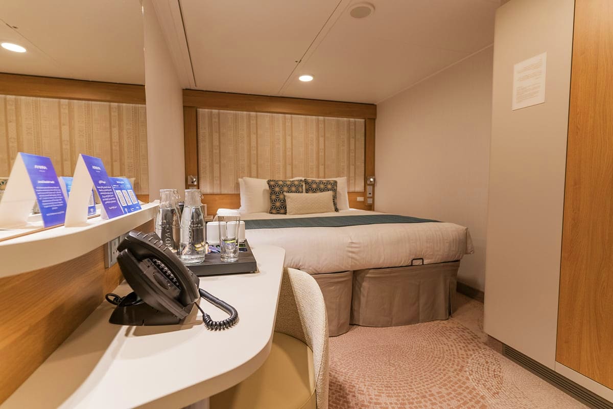 Modern cruise ship cabin with queen bed, desk with telephone, blue information cards, water glasses, and neutral beige and wo