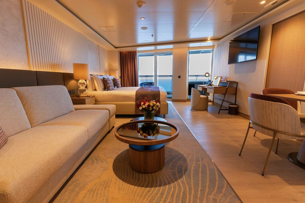 Luxury cruise ship suite with beige sofa, round center table with flowers, bed, desk, and ocean view balcony
