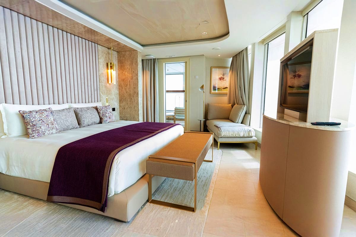 Luxury cruise ship bedroom with king bed, purple throw blanket, ocean view windows, sitting area, and modern beige and wood f