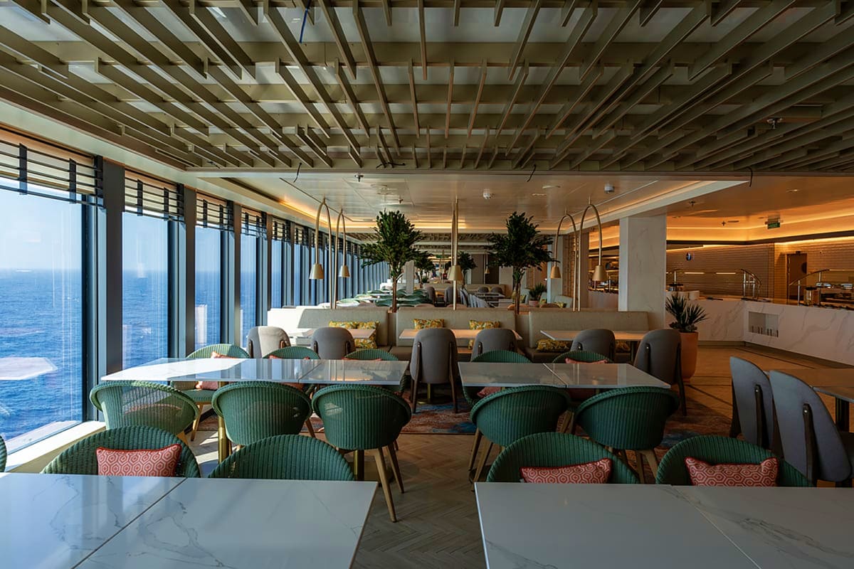 Modern cruise ship dining venue with floor-to-ceiling ocean views, green dining chairs, marble tables, and contemporary ceili
