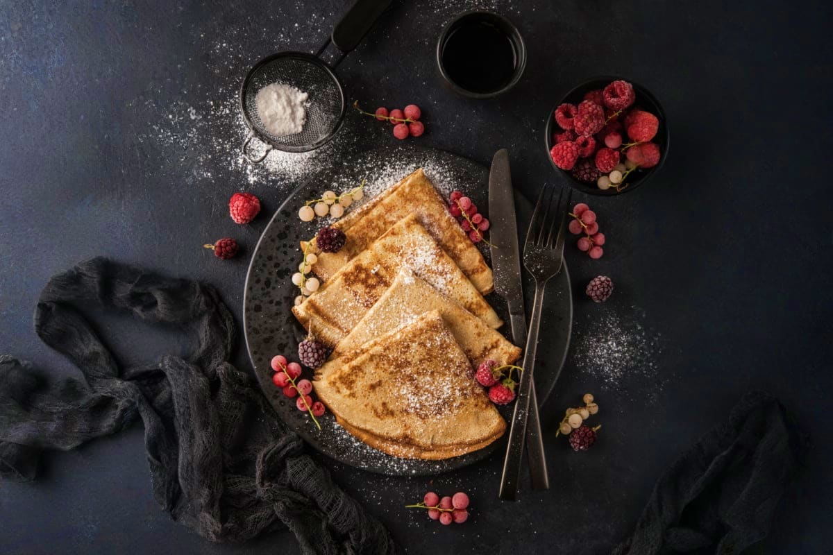 Crepes dusted with powdered sugar served with fresh raspberries and currants on a dark surface
