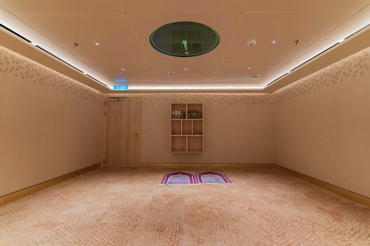 Modern prayer room on Aroya Aqaba cruise ship with carpeted floor, wooden door, shelving, and centered prayer mats