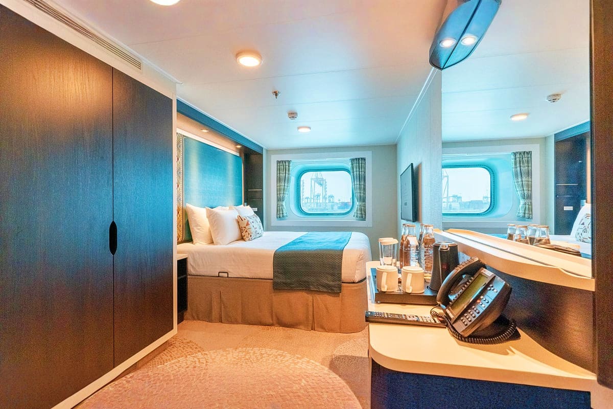 Modern cruise ship cabin with queen bed, two porthole windows overlooking ocean, desk with phone, and wood and blue interior 