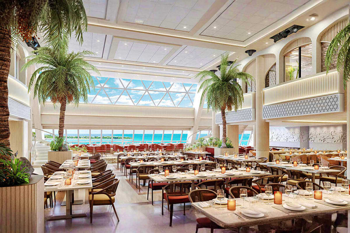 Modern cruise ship dining room with ocean views, palm trees, and tables set for service overlooking turquoise water.