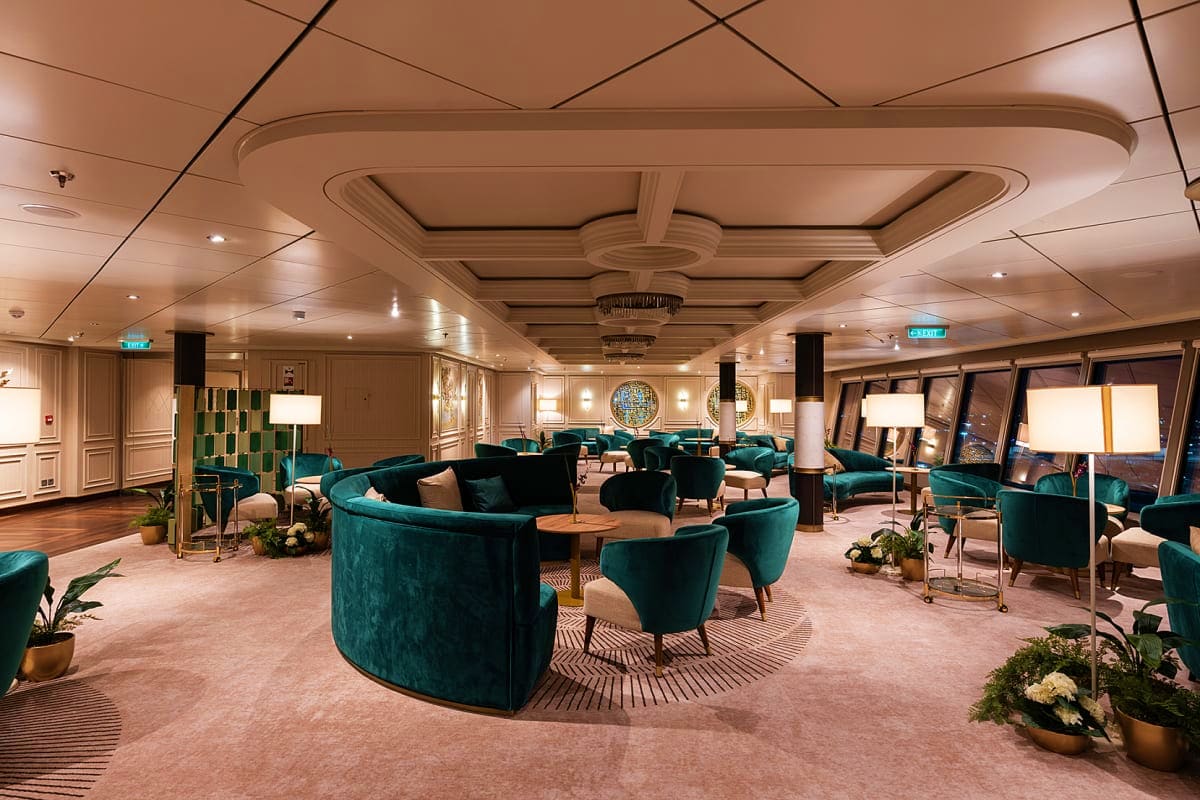 Elegant cruise ship lounge with teal velvet seating, coffered ceiling, decorative plants, and ocean views through large windo
