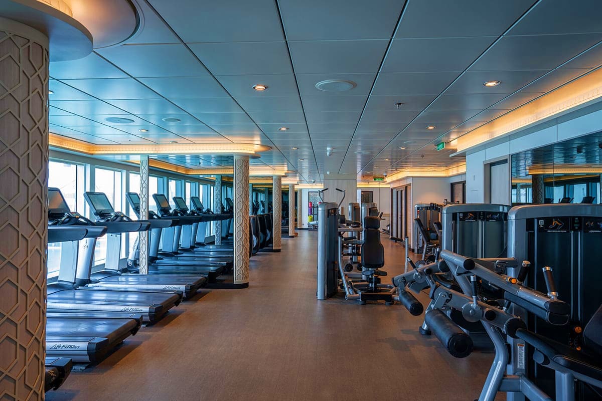 Modern cruise ship fitness center with treadmills, exercise equipment, and ocean view windows under illuminated ceiling
