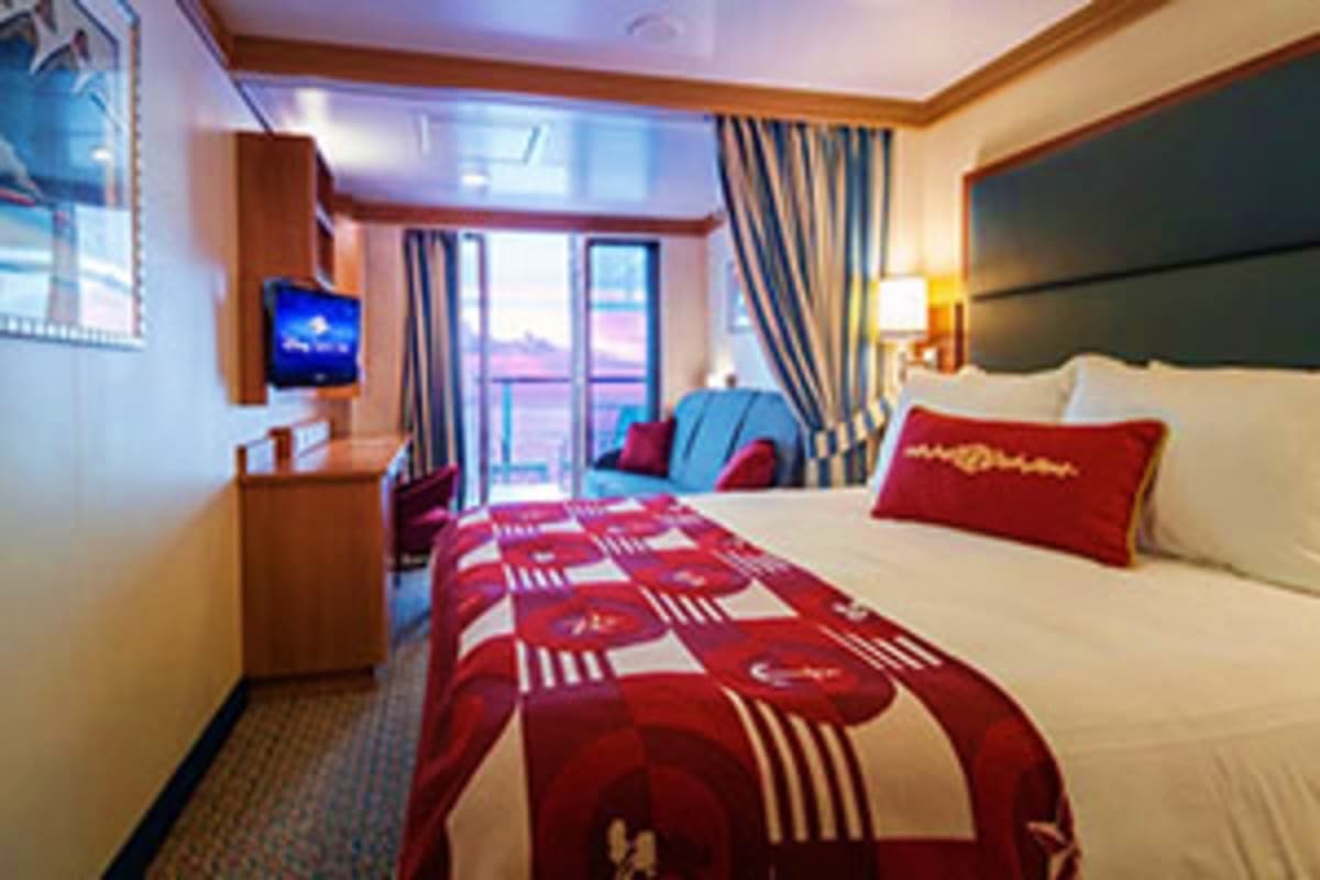 Spacious cruise ship cabin with queen bed, balcony view, flat-screen TV, and sitting area with blue and red accent pillows