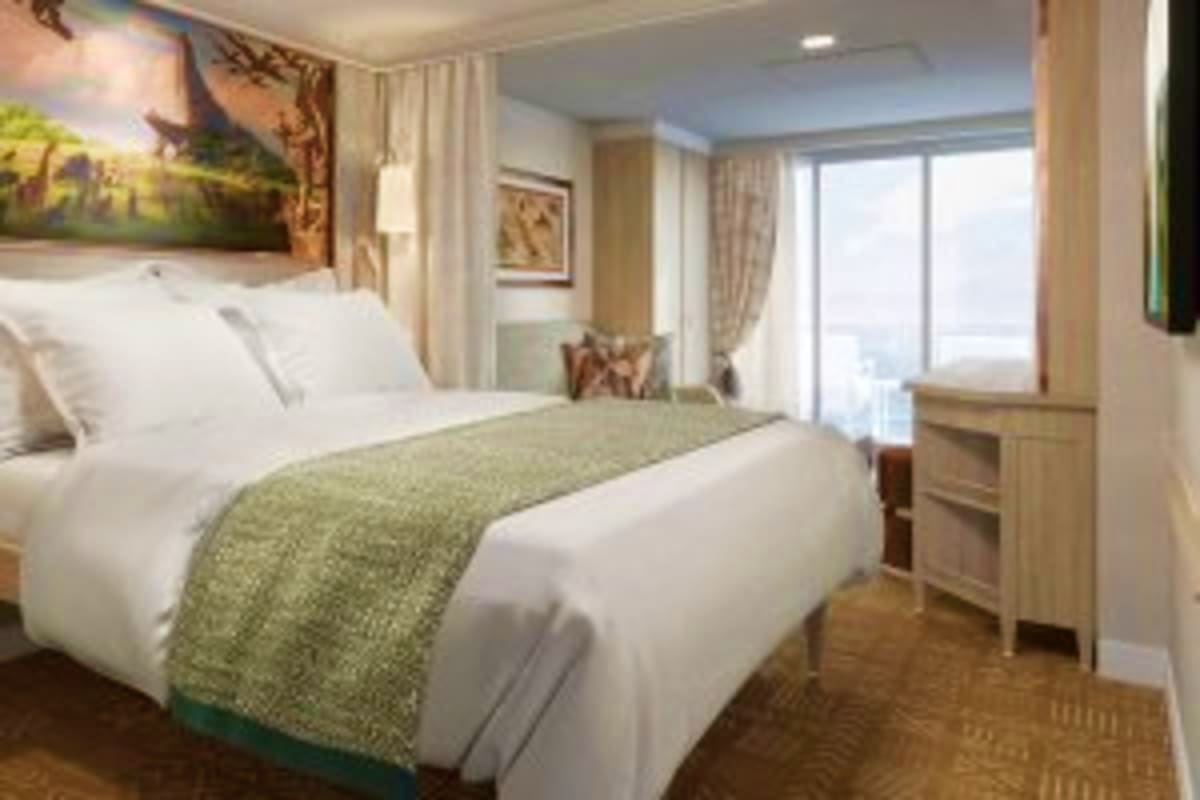 Modern cruise ship stateroom with queen bed, white bedding, green throw blanket, wood nightstand, ocean view balcony, and lan
