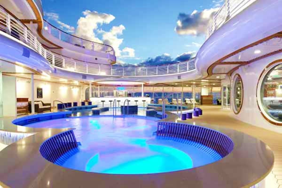 Luxury cruise ship indoor pool deck with illuminated blue pools, curved architecture, ocean views, and sky visible through gl
