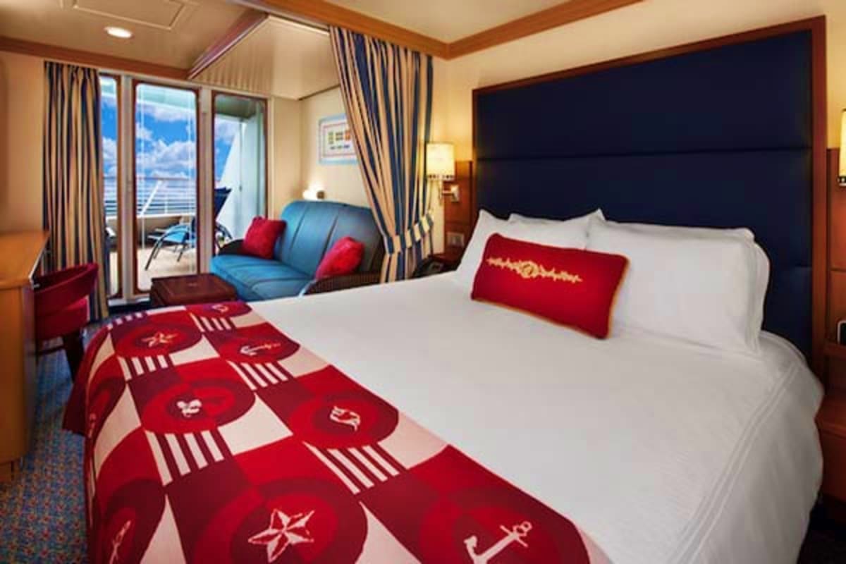 Luxurious cruise ship stateroom with queen bed, balcony overlooking ocean, blue sofa with red pillows, and nautical-themed re