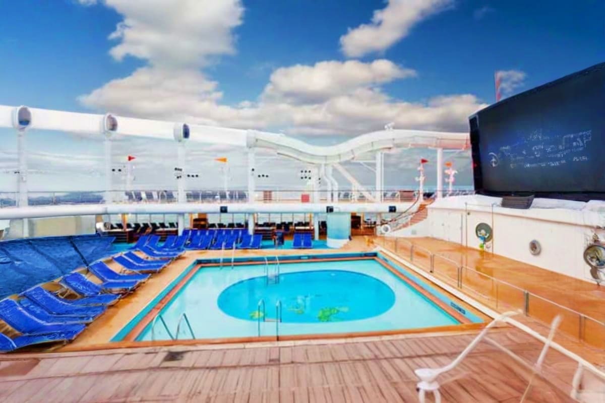 Cruise ship pool deck with bright blue lounge chairs, oval swimming pool, white ship superstructure, and open sky above