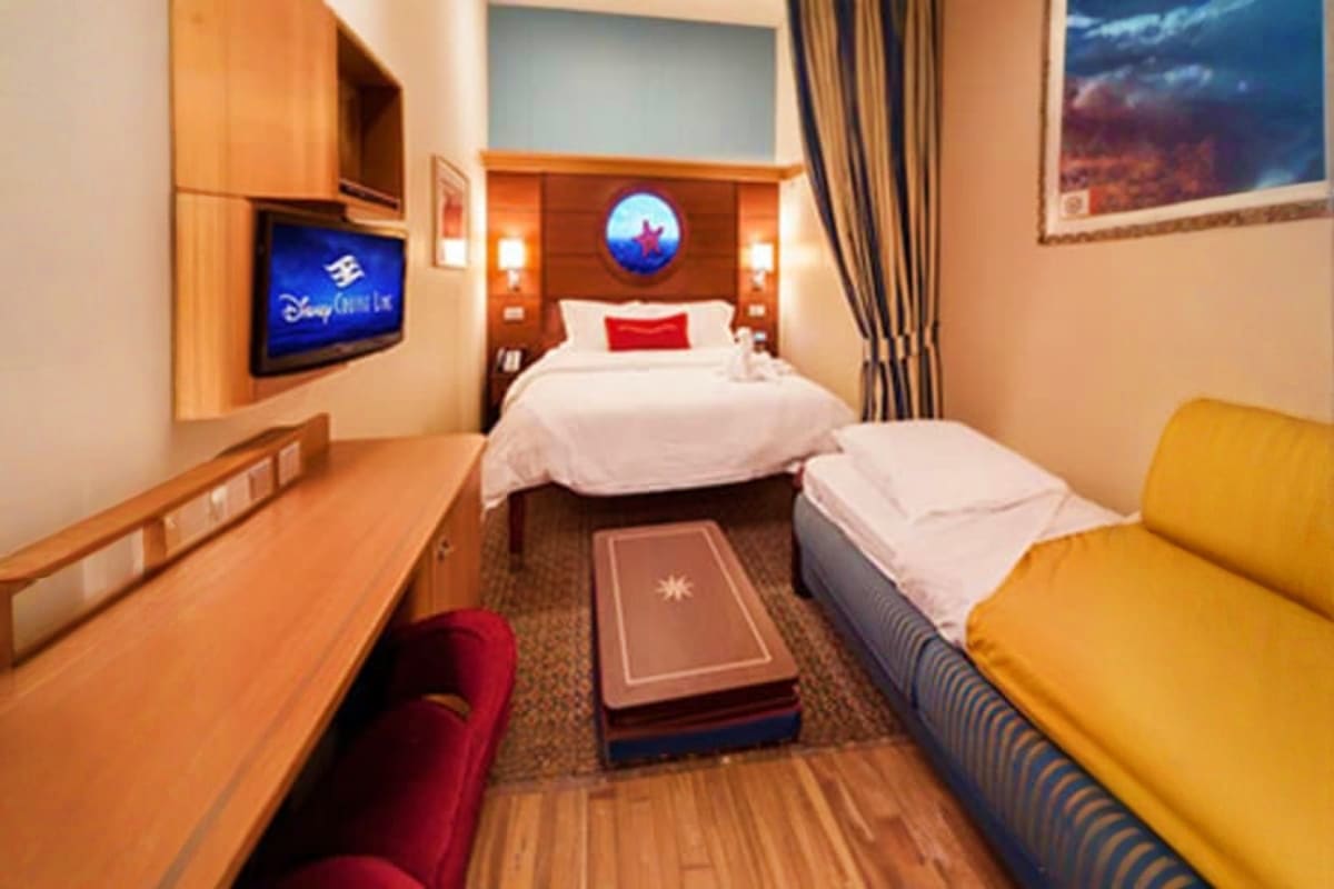 Disney-themed cruise ship cabin with queen bed, twin bed, TV, desk, and yellow chair seating area