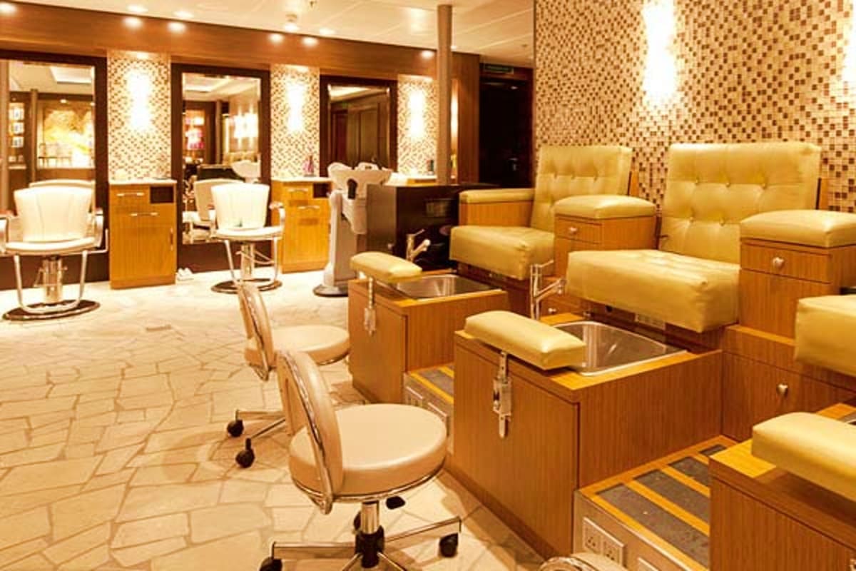 Luxury cruise ship spa with pedicure stations, golden cushioned chairs, wooden cabinetry, and mosaic tile walls with ambient 