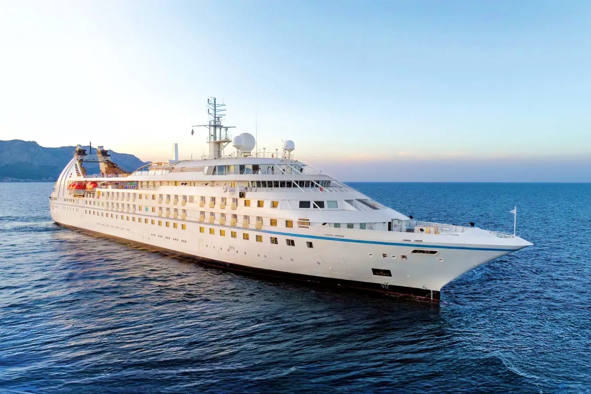 Large luxury cruise ship with white hull and multiple decks anchored in blue ocean waters with coastal mountains visible in b