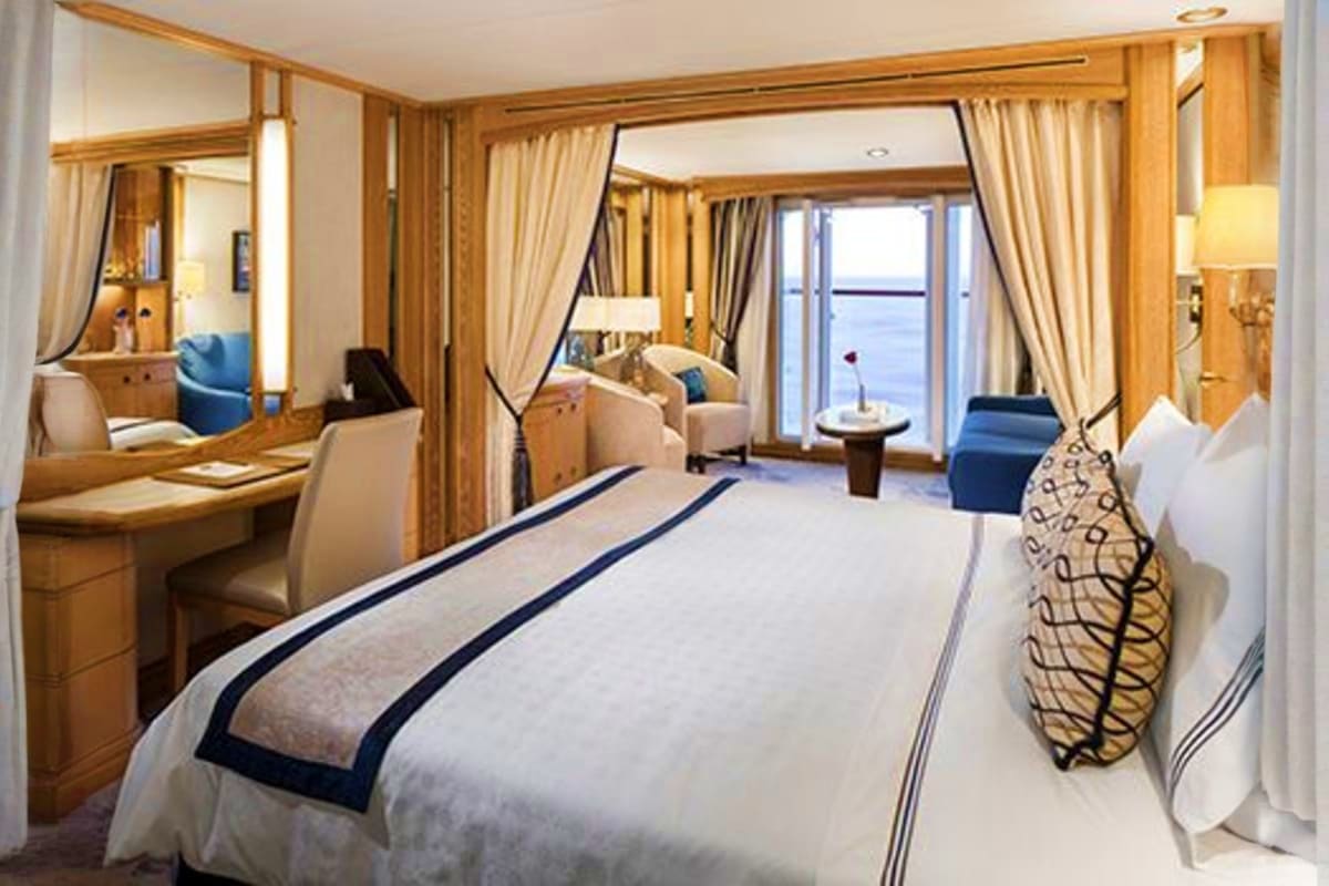 Luxury cruise ship cabin with queen bed, wooden furnishings, ocean view balcony, and sitting area with blue accents