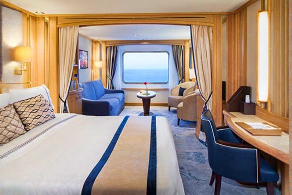 Luxury cruise ship cabin with ocean view window, bed, blue armchair, seating area, and natural light from large porthole wind