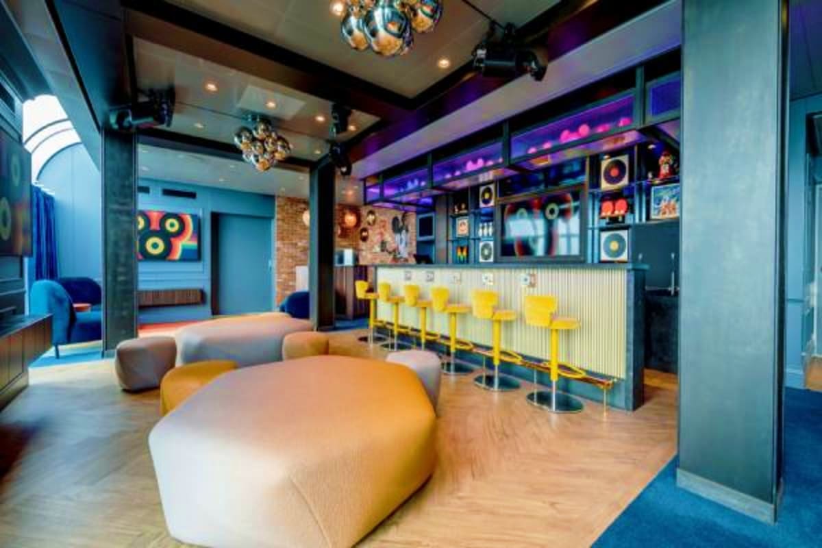 Modern lounge with yellow bar stools, curved seating, colorful speaker wall displays, and blue neon accents in cruise ship en