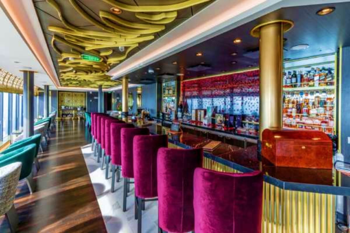 Modern cruise ship bar interior with burgundy velvet bar stools, gold ceiling details, and shelves of liquor bottles