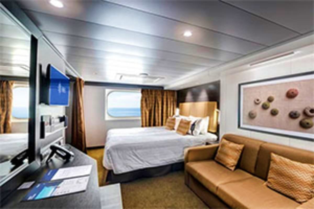 Luxury cruise ship stateroom with queen bed, tan sofa, flat-screen TV, porthole windows with ocean view, and modern decor