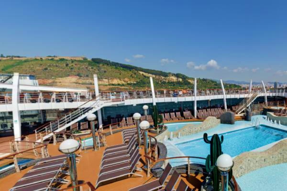 Cruise ship deck with swimming pool, striped lounge chairs, and hillside landscape in background under clear blue sky