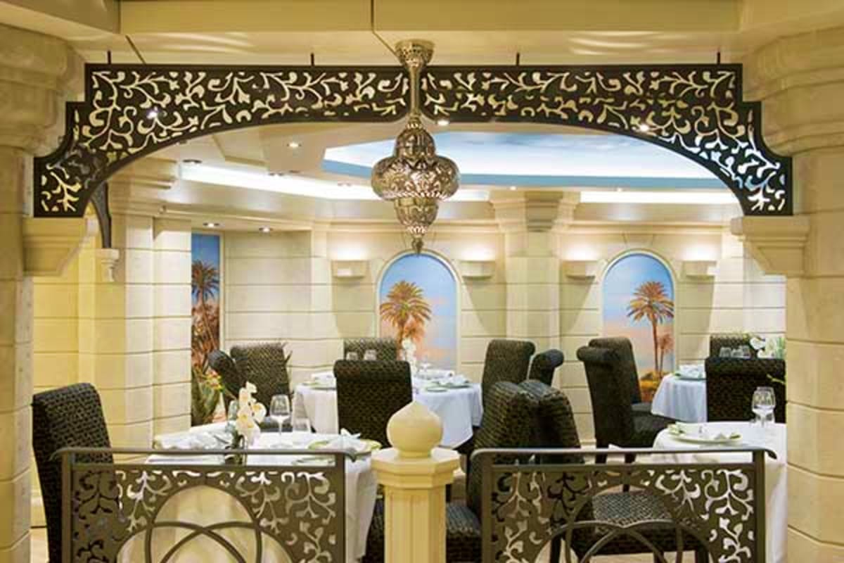 Elegant fine dining restaurant interior with ornate brass railings, white tablecloths, palm tree murals, and crystal chandeli