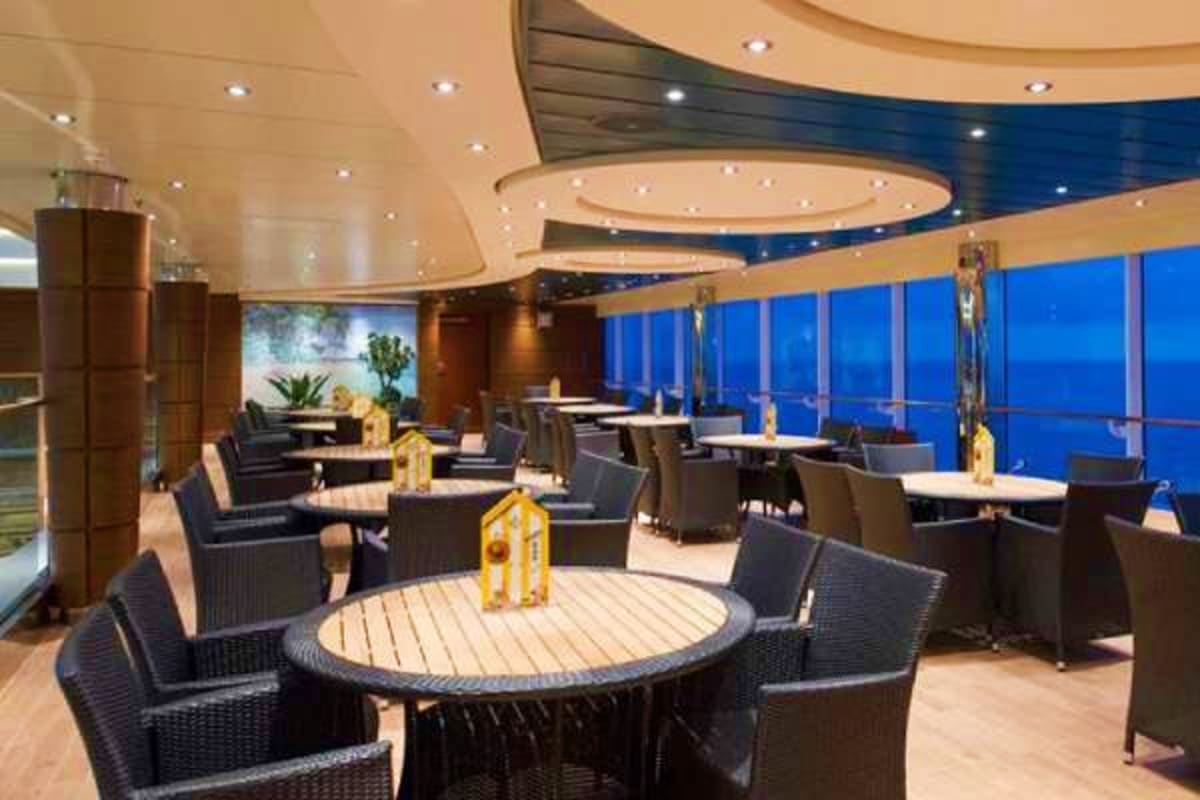 Upscale cruise ship dining venue with round tables, black chairs, floor-to-ceiling ocean views, and modern ceiling design