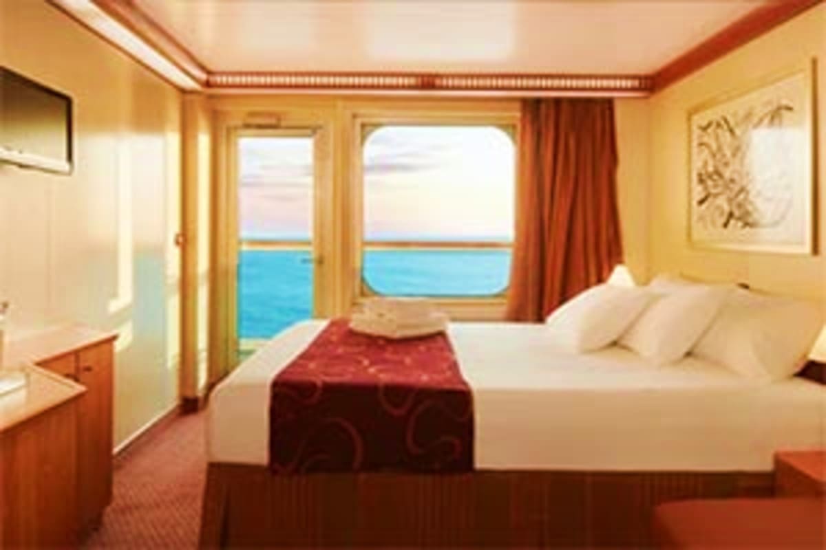 Luxury cruise ship cabin with ocean view balcony, queen bed with burgundy runner, and floor-to-ceiling windows overlooking bl