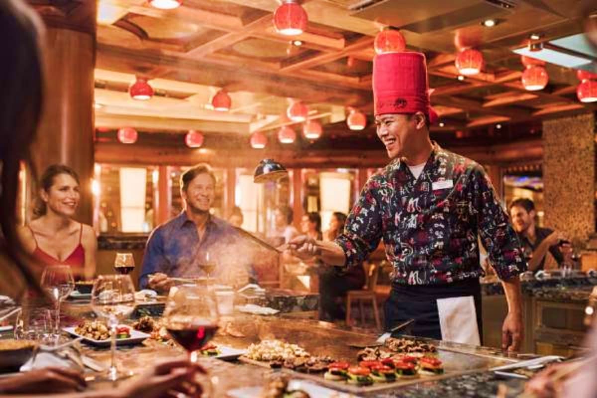 Chef in tall red hat entertaining guests at open kitchen counter during dinner service on Costa Fascinosa cruise ship