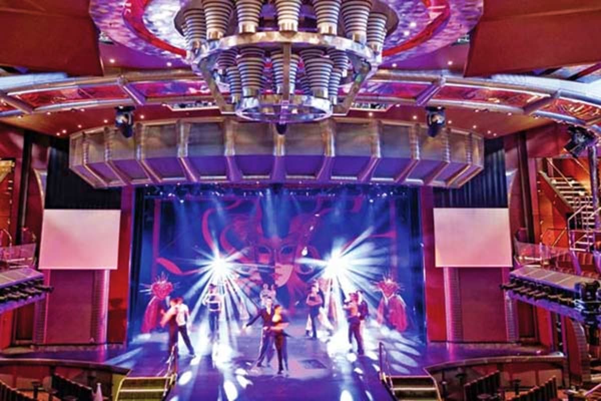 Modern cruise ship theater with red and purple lighting, performers on stage, and dramatic spotlights illuminating the entert