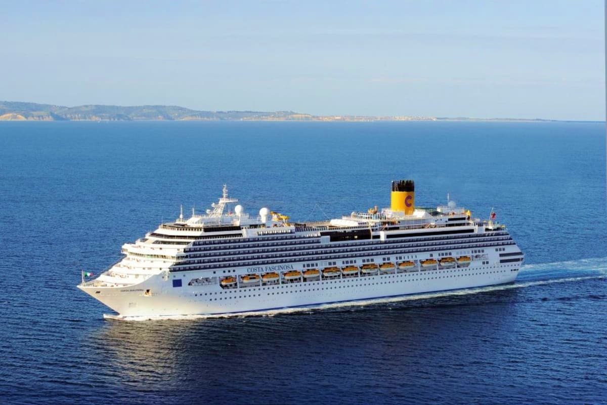 Large white Costa cruise ship with yellow funnel sailing on blue Mediterranean sea with coastal cliffs in background