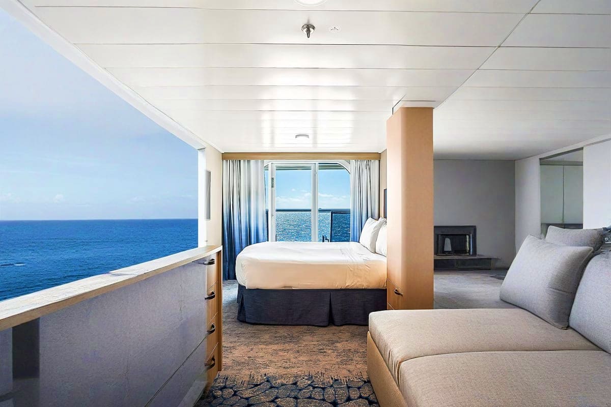 Luxury cruise ship cabin with ocean view balcony, queen bed, sofa, and floor-to-ceiling windows overlooking blue sea