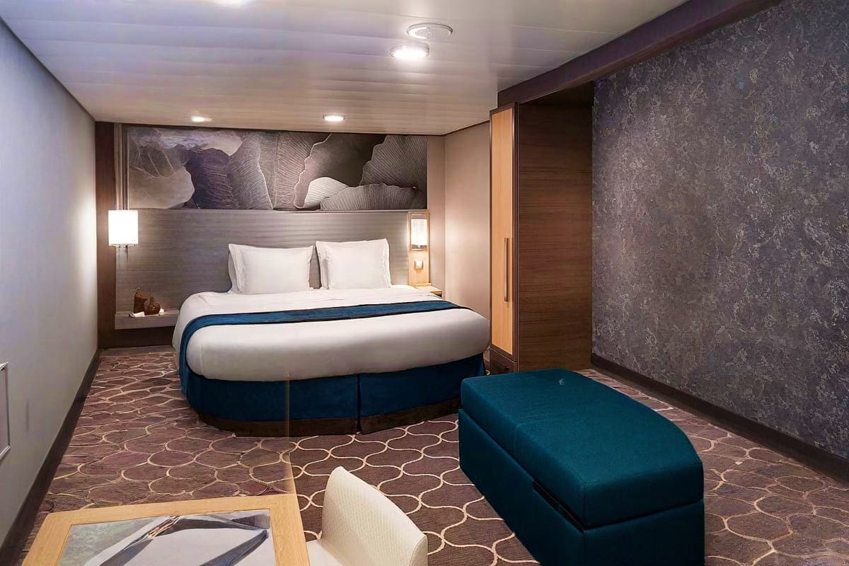 Modern cruise ship cabin with round bed, teal ottoman seating, dark wood paneling, and abstract headboard artwork
