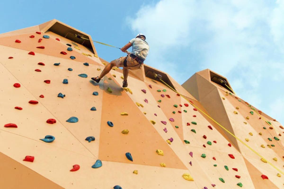Person rock climbing on an outdoor climbing wall with colorful handholds under blue sky
