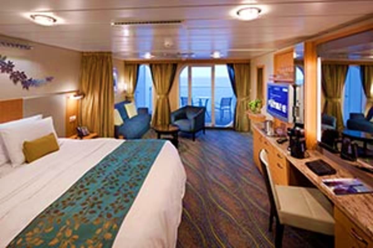 Luxury cruise ship stateroom with king bed, seating area, balcony with ocean view, and modern wood furnishings