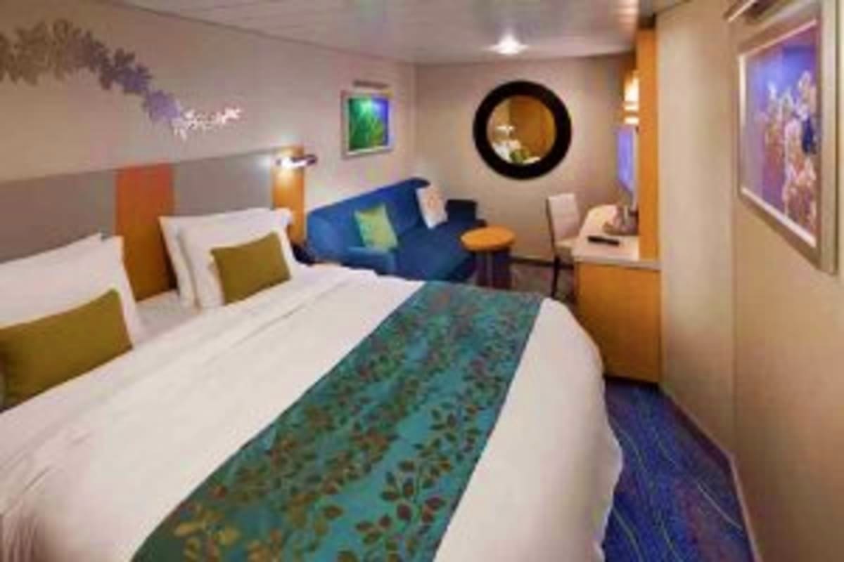 Modern cruise ship cabin with queen bed, blue sofa, round porthole window, and contemporary décor in neutral and ocean blue t