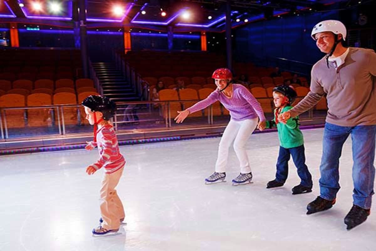 Adult and children ice skating together on an indoor ice rink with blue and purple lighting and orange stadium seating