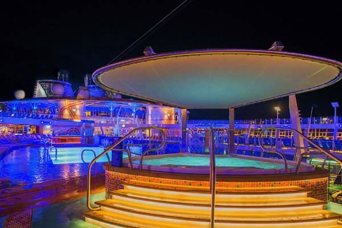 Night view of cruise ship deck with illuminated hot tub, swimming pool, and lounge seating under starry sky