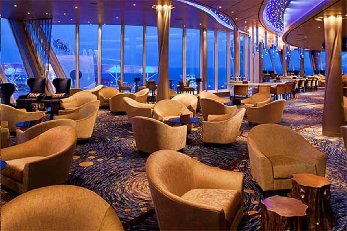 Elegant lounge area on Vision of the Seas with beige chairs, ocean view through floor-to-ceiling windows at dusk, and modern 