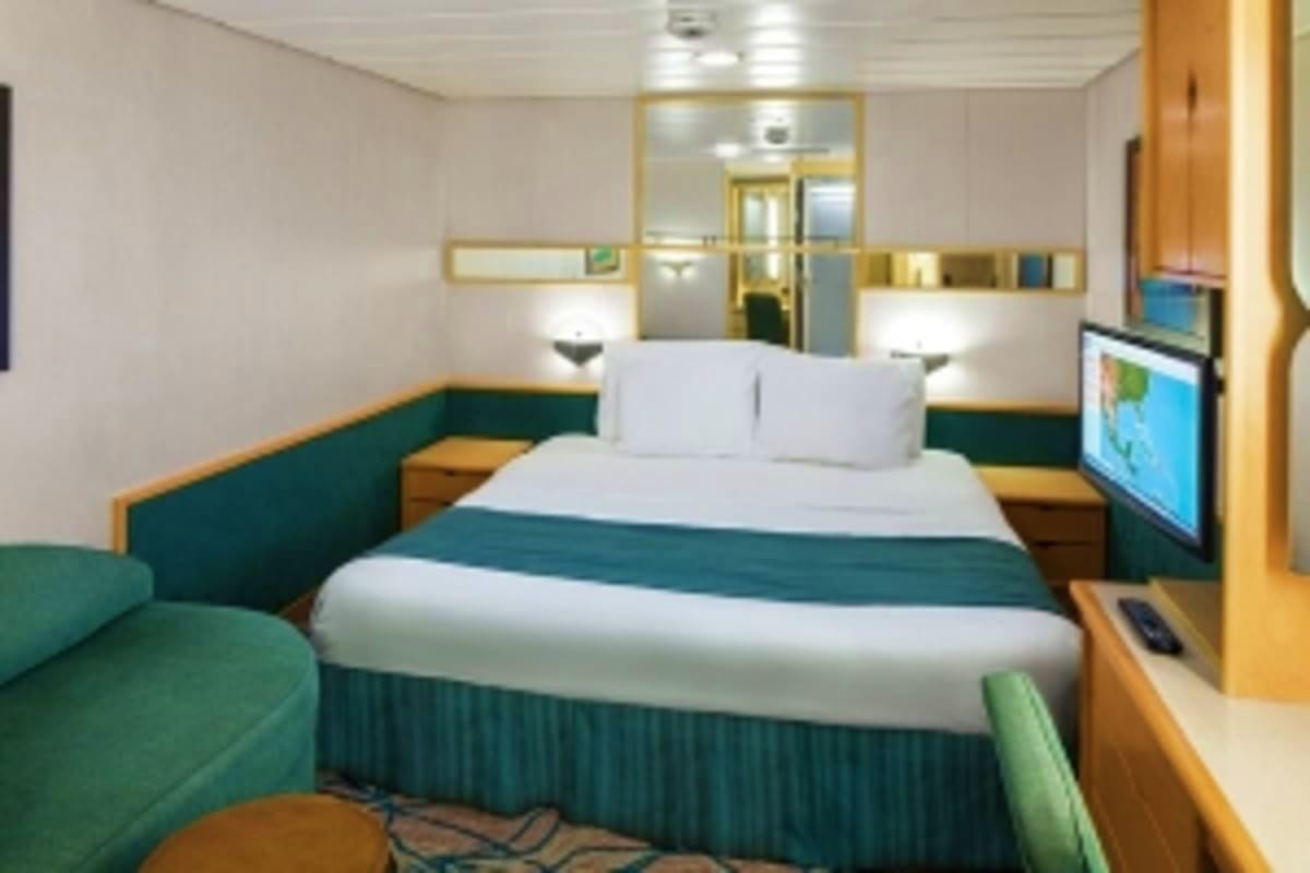 Modern cruise ship stateroom with queen bed, green upholstered seating, flat-screen TV, and mirrored vanity area
