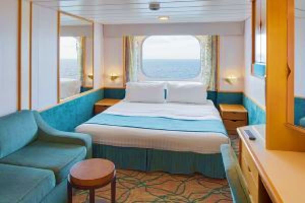 Cruise ship cabin with ocean view window, queen bed, turquoise seating area, and natural lighting