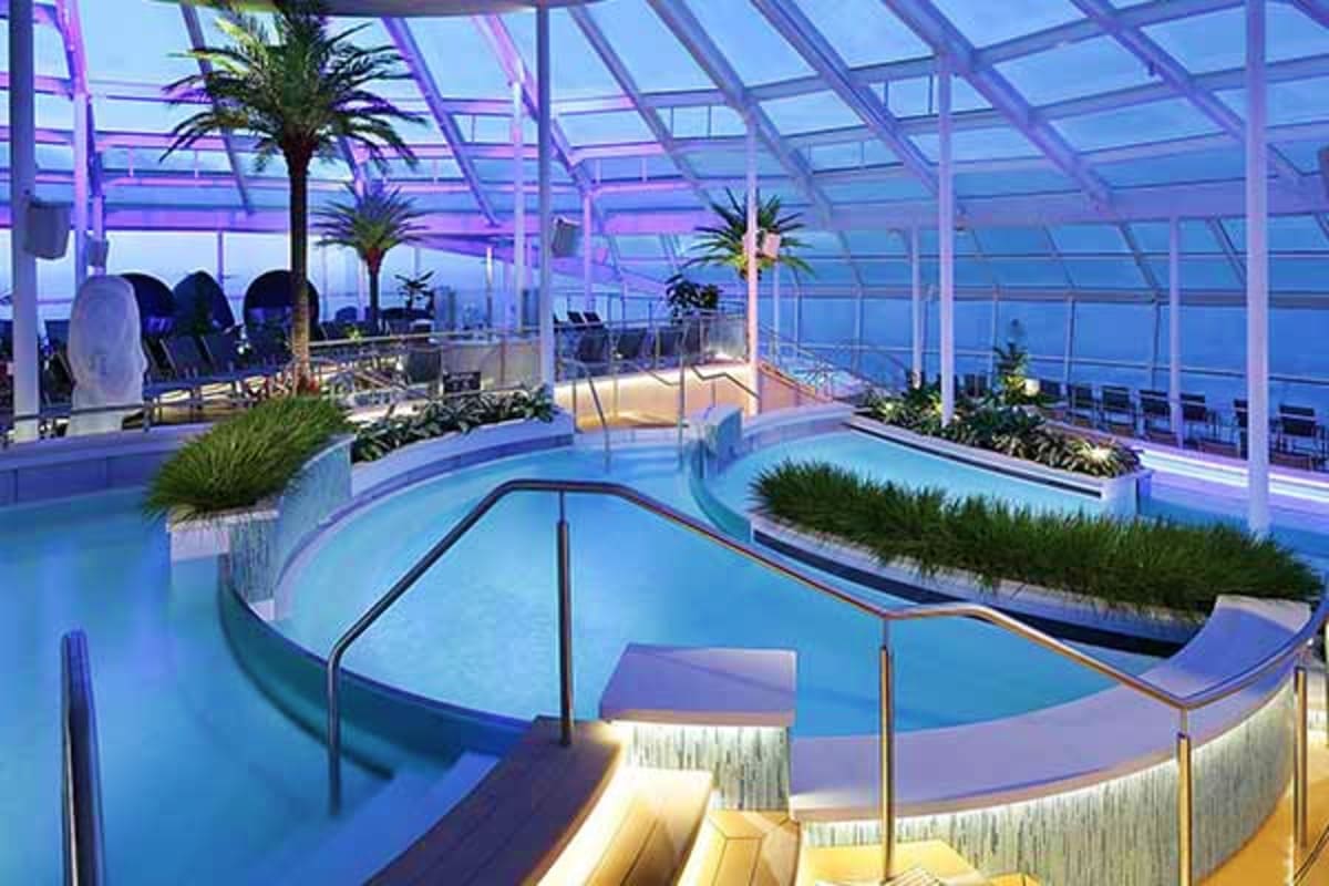 Modern cruise ship indoor pool area with curved turquoise pools, palm trees, glass ceiling with purple lighting, and ocean vi