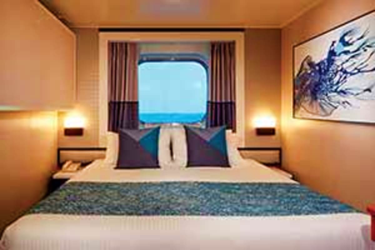 Norwegian Jade Japan Cruise Cruise ship cabin with ocean view window, bed with decorative pillows, warm lighting, and blue sea visible through porthole w