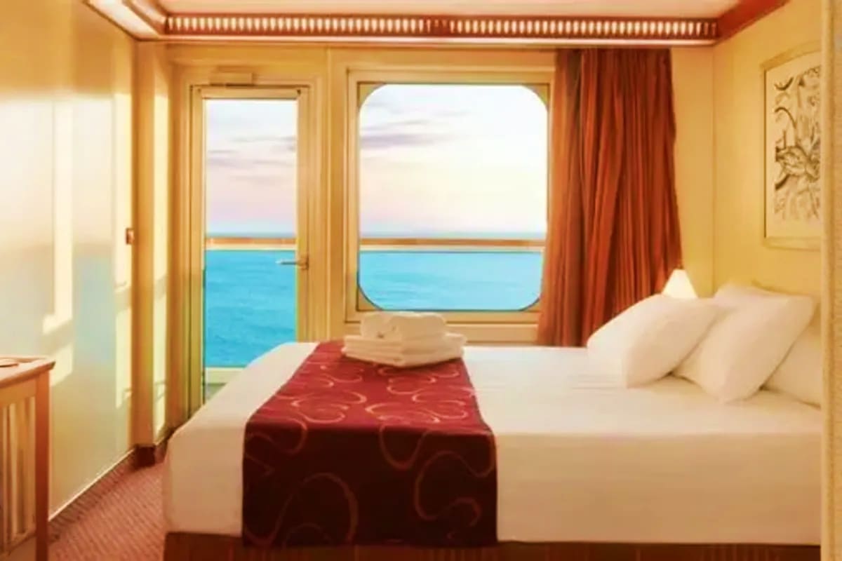Luxury cruise ship balcony cabin with ocean view, queen bed with burgundy throw, cream pillows, and glass doors opening to se