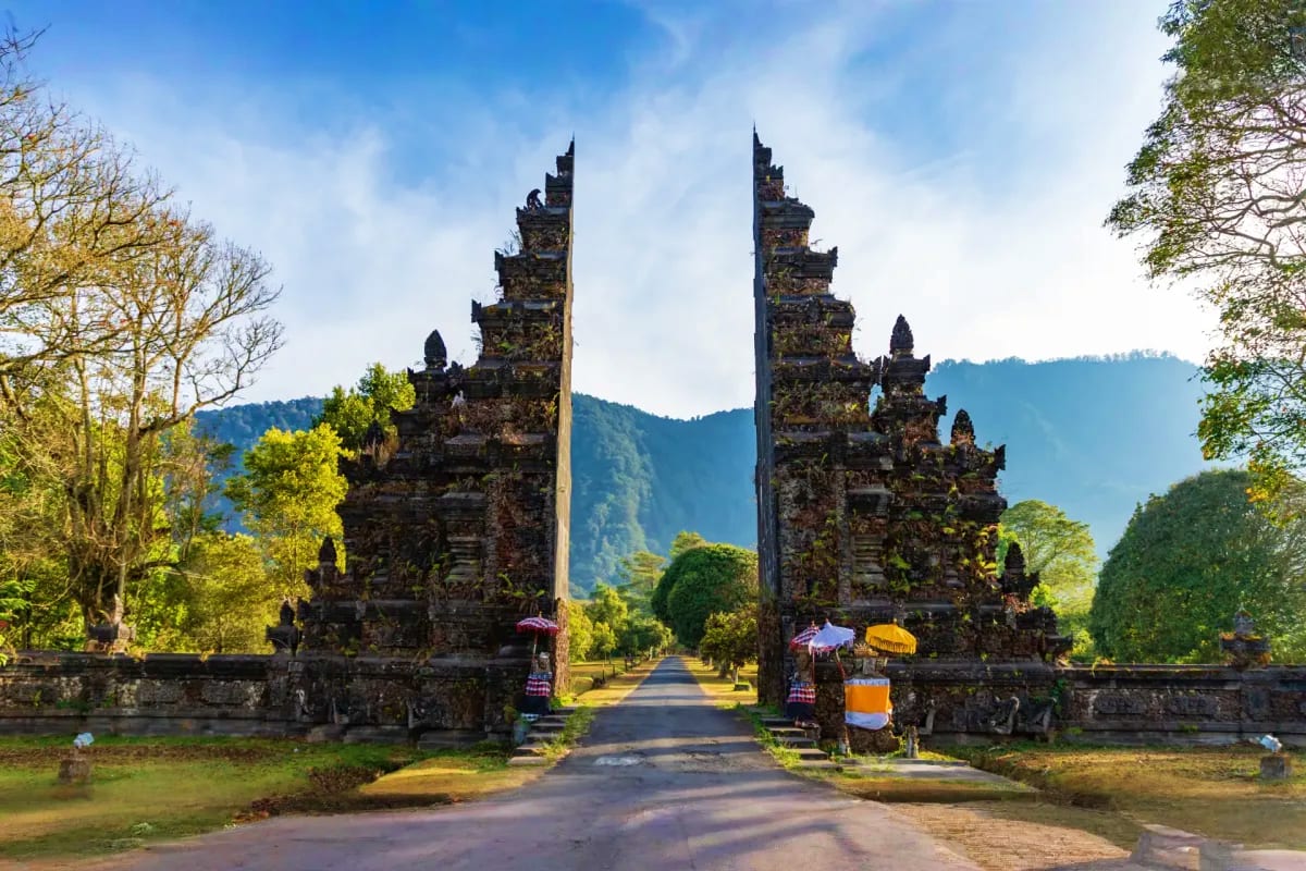 Twin ornate stone temple gates with carved spires flanking a tree-lined pathway in lush Balinese landscape with forested moun
