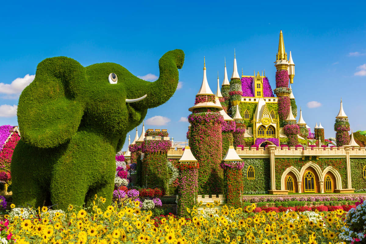Whimsical topiary elephant and ornate castle covered in flowering vines and colorful blooms against blue sky