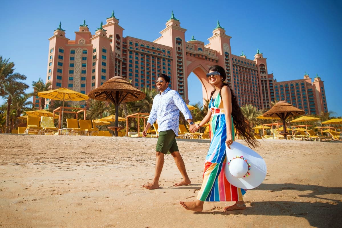 Couple walking on sandy beach in front of Atlantis The Palm luxury hotel in Dubai with yellow umbrellas and palm trees