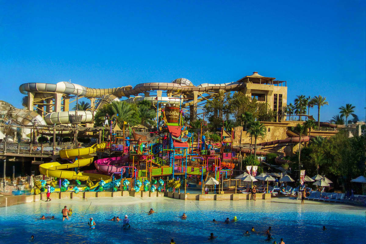 Colorful water park with large yellow and pink water slides, swimming pool with visitors, palm trees, and tan-colored buildin